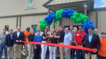 Domtar celebrated the EAM expansion in Jesup, GA on May 24. Pictured: Group of people in front of green and blue balloon arch and EAM facility exterior, with 3 people in the middle holding scissors to cut the ribbon on the new expansion. The ribbon is red-orange and being held by two men about 12 feet apart.