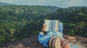 Image: man in straw hat, laying on mountaintop overlooking green mountains and reading a book. Check out this list of nine books by Indigenous authors. Newsroom.Domtar.com.