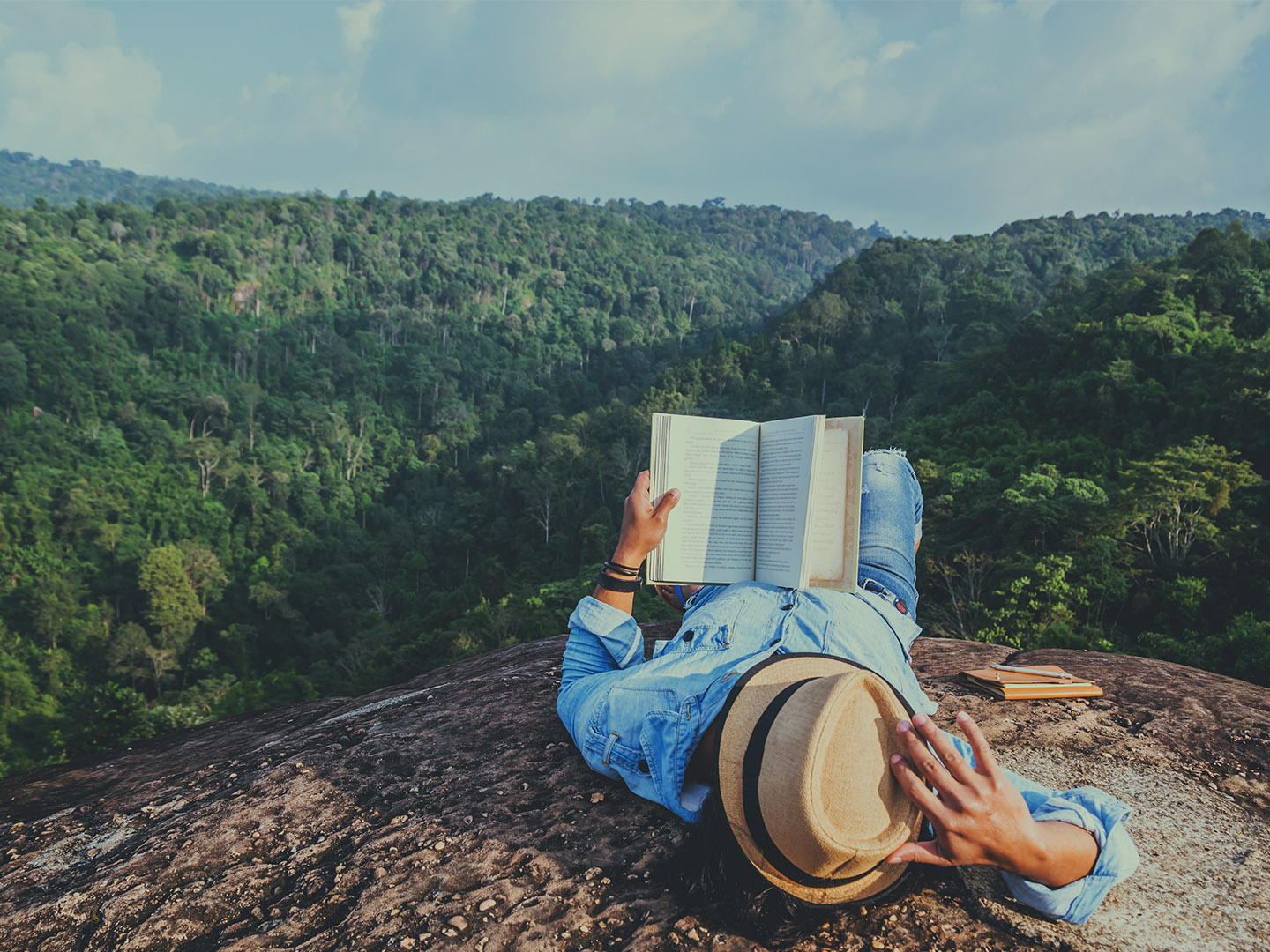 Image: man in straw hat, laying on mountaintop overlooking green mountains and reading a book. Check out this list of nine books by Indigenous authors. Newsroom.Domtar.com.