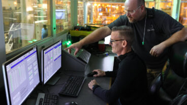 Two male workers in a control room at a pulp mill are looking at a computer screen. The man in the back is standing and pointing something out on the screen to the other man, who is seated. Institutional knowledge is being shared to help each other do their jobs.
