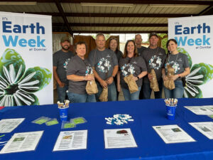 Ashdown Mill EarthChoice Ambassadors hosted a seedling giveaway - employees are pictured with Earth Week banners and a blue tablecloth-covered table with additional info sheets.