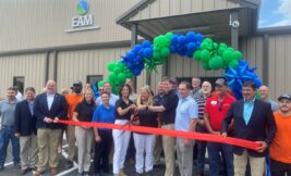 Domtar celebrated the EAM expansion in Jesup, GA on May 24. Pictured: Group of people in front of green and blue balloon arch and EAM facility exterior, with 3 people in the middle holding scissors to cut the ribbon on the new expansion. The ribbon is red-orange and being held by two men about 12 feet apart.