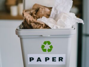 Image: Grey recycling bin in office environment, labeled "PAPER" with the recycling symbol. There are brown and white crumpled papers coming out of the top. Domtar continues to support higher recycling rates of paper and cardboard.